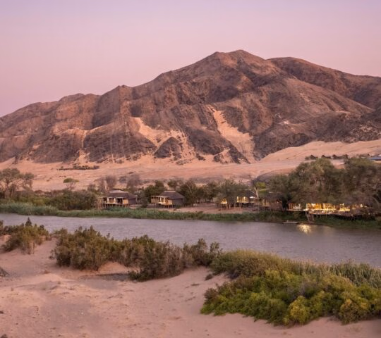 Aerial view of Wilderness Serra Cafema lodge on the banks of a river surrounded by desert dunes and rocky mountains.