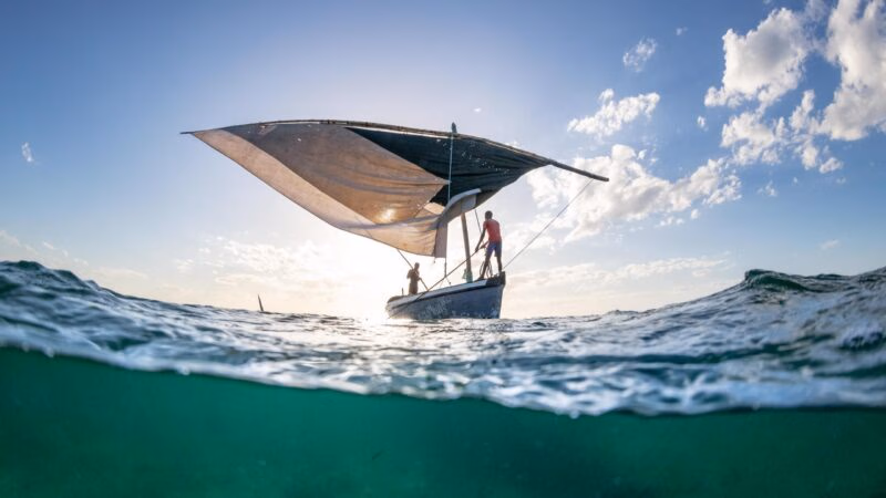A split underwater and surface view of a traditional dhow boat sailing on clear turquoise ocean waters.