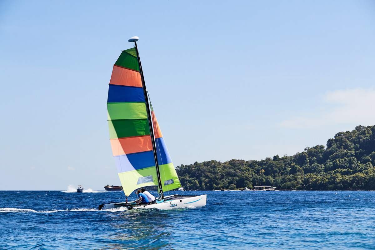 A colourful Hobie Cat catamaran sails on the ocean with a lush green island in the background.