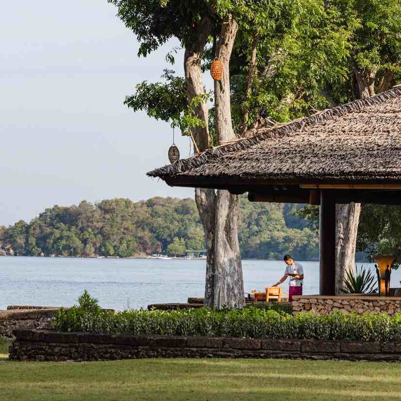 A man sets a table under a thatched roof structure next to a quiet ocean bay with forested hills.