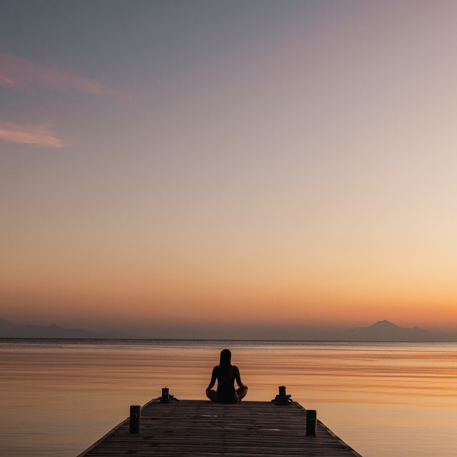 Silhouette of a person practicing yoga on a wooden pier at sunset over a still ocean.