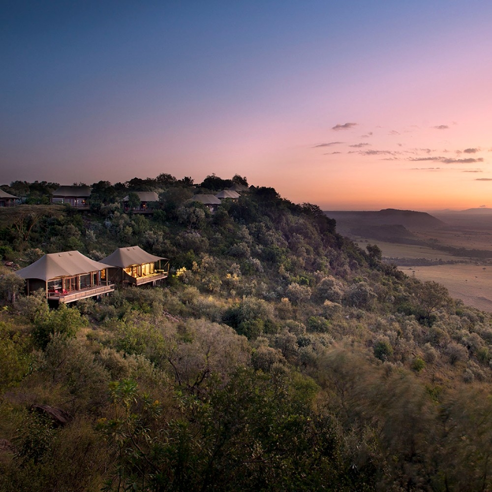 Luxury safari tents on a tree-covered ridge overlooking a wide valley at sunset with a pink and purple sky.