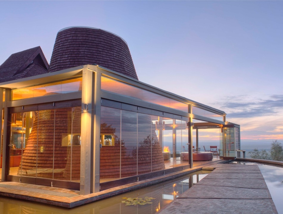 Modern glass-walled building with conical wooden structures inside, reflecting a sunset next to a water feature.