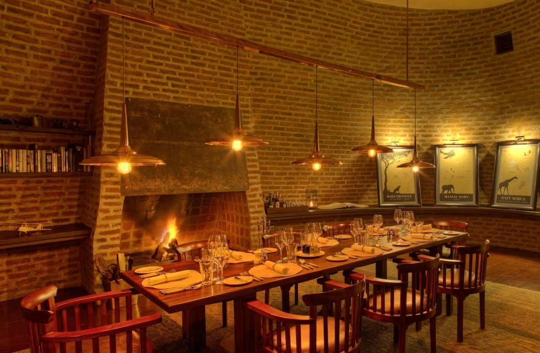 A formal wooden dining table set with glassware under hanging lamps in a circular brick room with a fireplace.