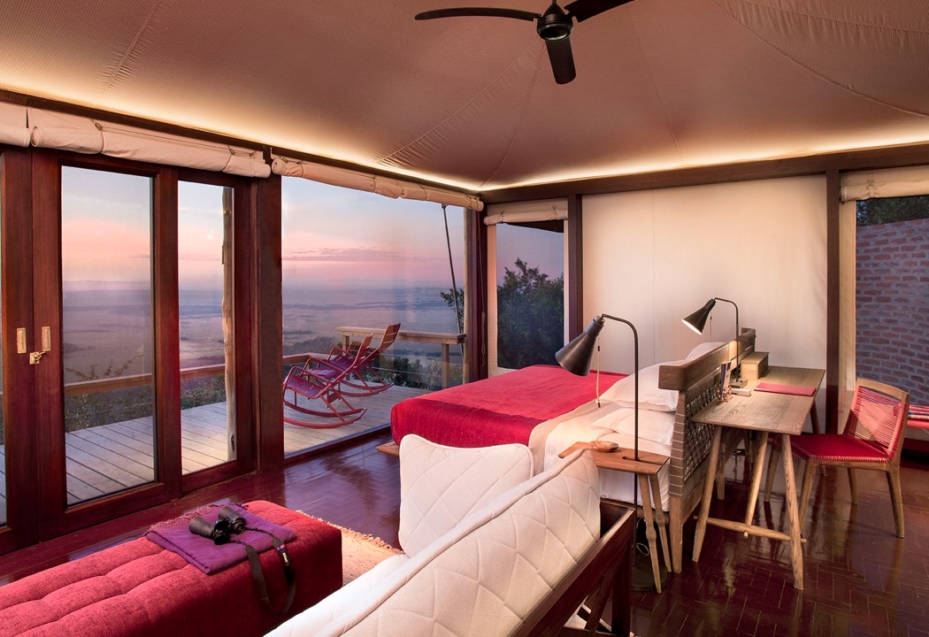 Interior of a tented suite with a red bedspread, wooden floors, and glass doors opening to a balcony and valley view.