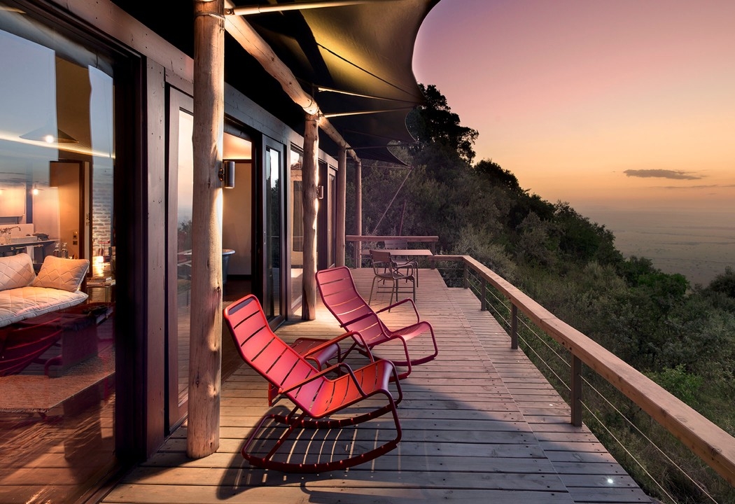 Two red rocking chairs on a wooden balcony overlooking a forested valley under a colorful sunset sky.