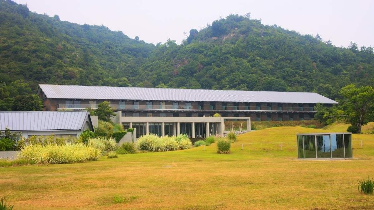 A long, two-story modern building with a grey roof situated at the base of a forested hill.