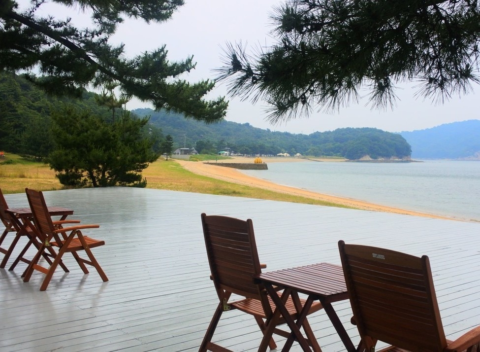 Wooden chairs and tables on a deck overlooking a beach and coastal hills under a hazy sky.