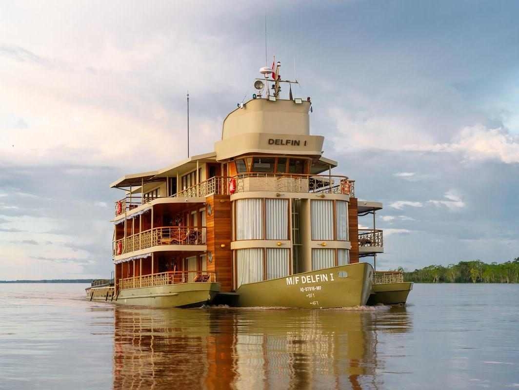 The exterior of Delfin I, a luxury wooden river cruise, sailing on a wide river under a cloudy sky.