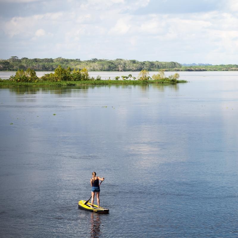 A woman paddleboarding on a wide, calm river with green trees and a cloudy sky in the distance.