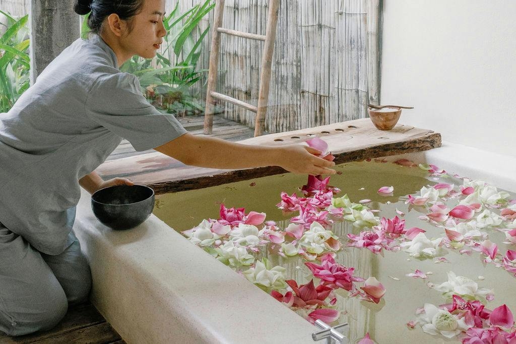 A person adding fresh pink lotus flowers to a white bathtub in a bright, natural spa setting.