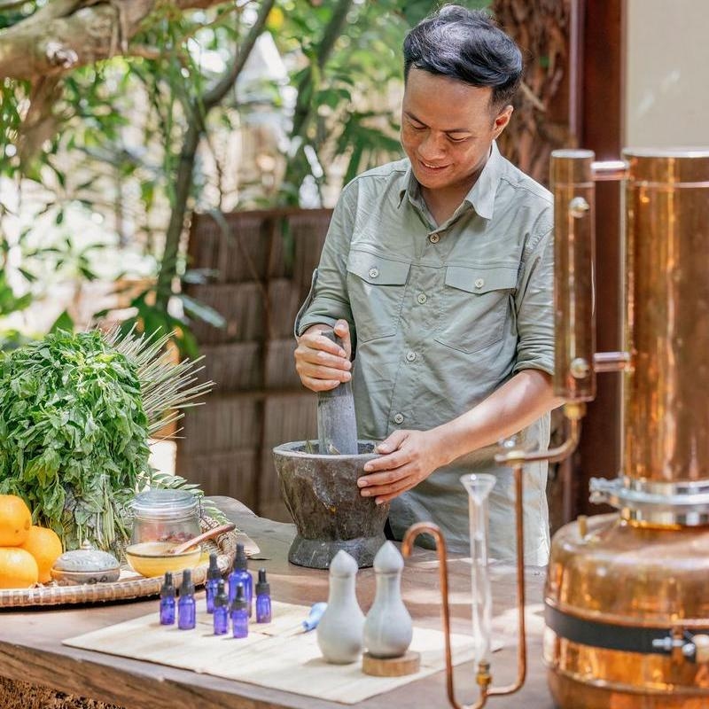 A person grinding herbs in a mortar and pestle next to blue glass bottles and copper spa equipment.