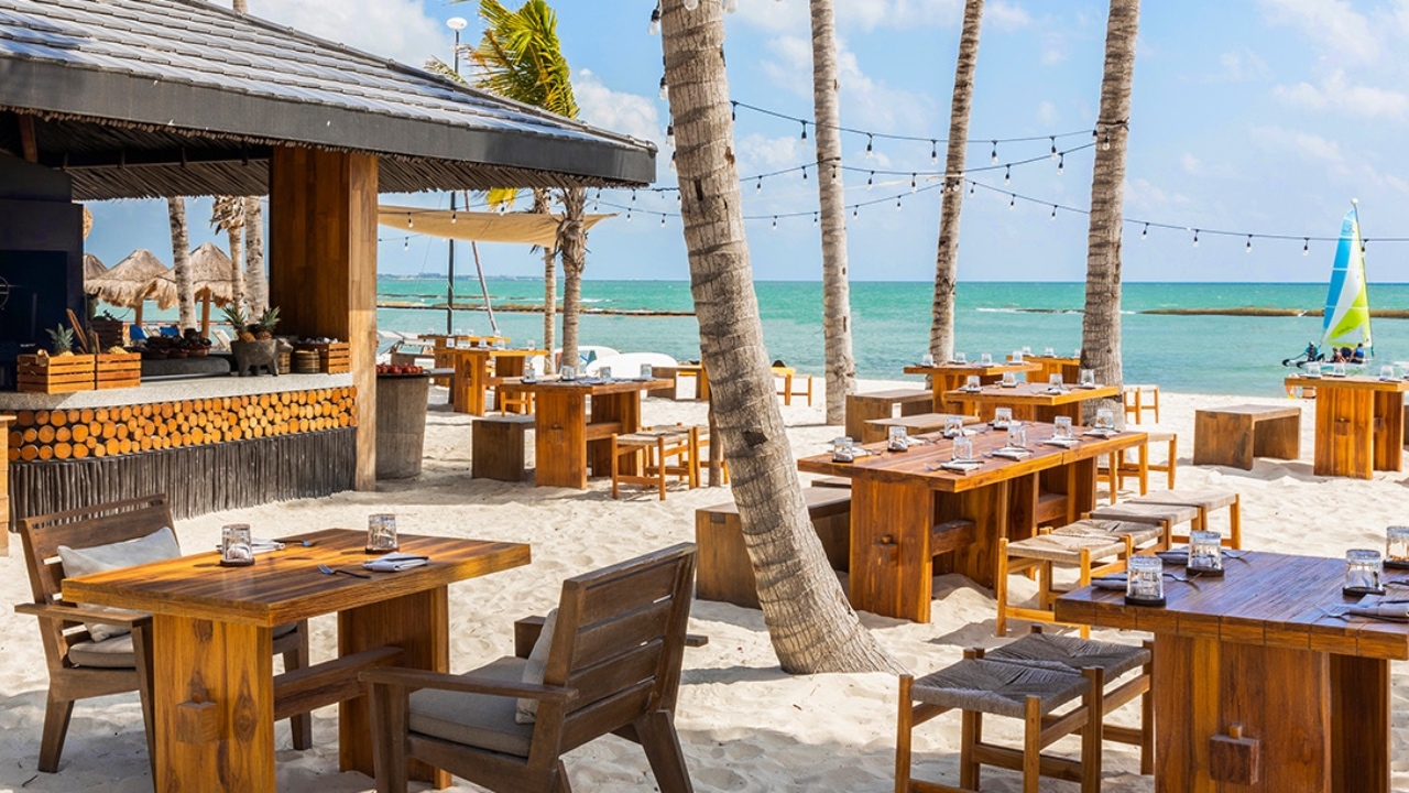 Beachfront dining area with wooden tables and chairs, string lights, and a clear blue sky.