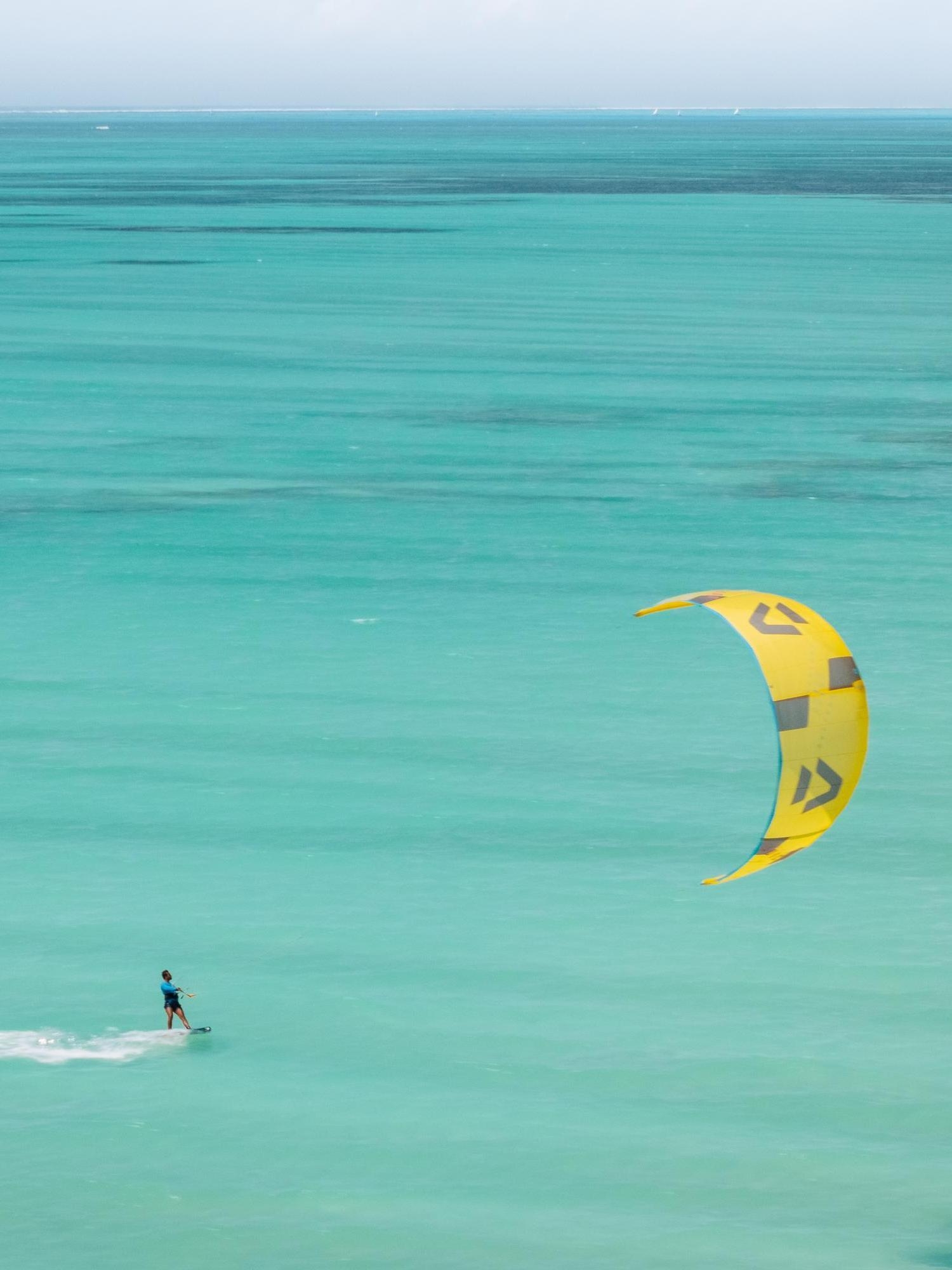 A person kiteboarding on the turquoise waters of a calm ocean, with a large yellow kite aloft.