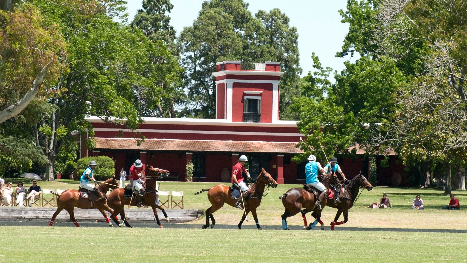 Polo players on horses in a field with a red and white structure in the background.