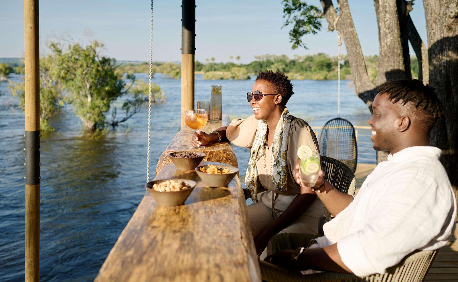 A man and woman relaxing with cocktails at a wooden bar overlooking the river at Victoria Falls River Lodge.