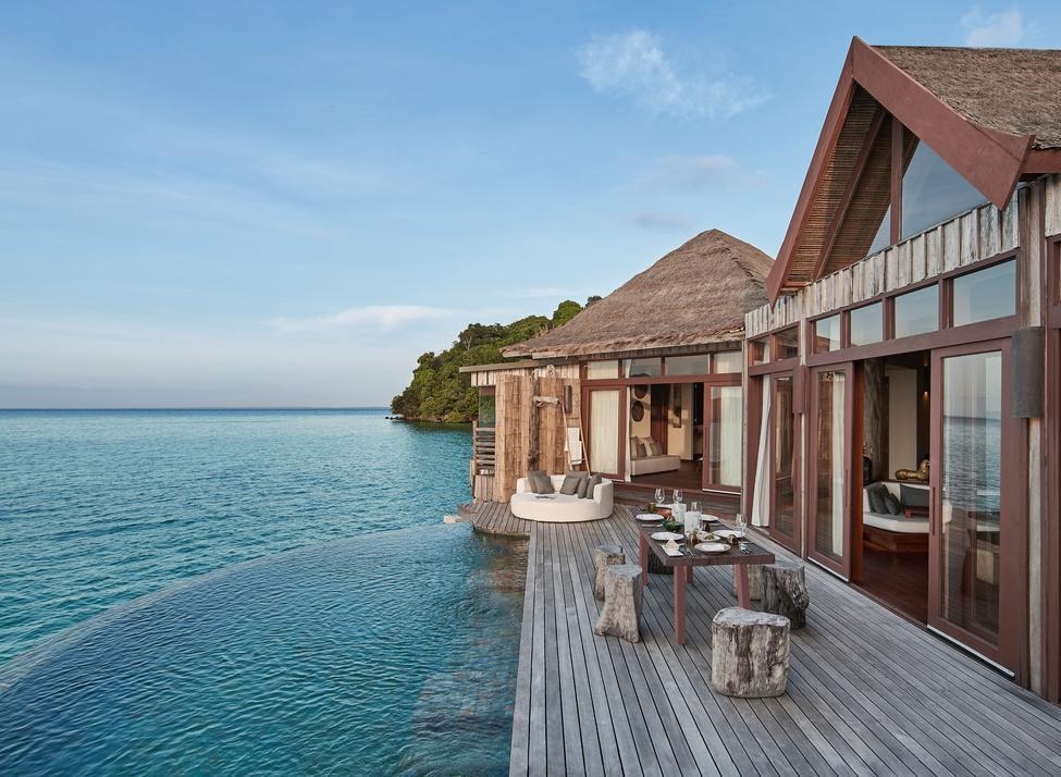 A wooden overwater villa with a private pool and outdoor dining table set against a clear blue sky.