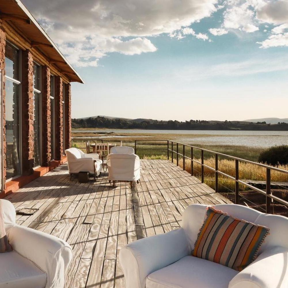 Wooden hotel terrace with white lounge chairs overlooking a calm lake and grassy wetlands under a bright sky.
