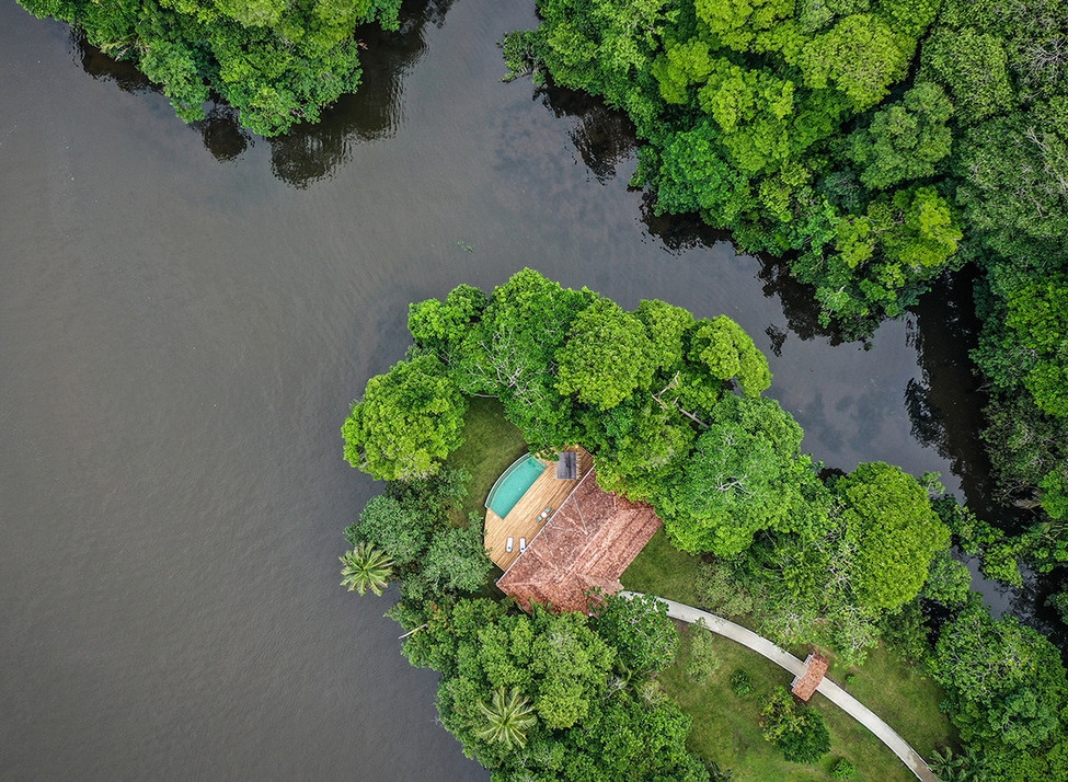 Aerial view of a luxury villa with a pool and red roof nestled in thick green jungle foliage beside a dark river.
