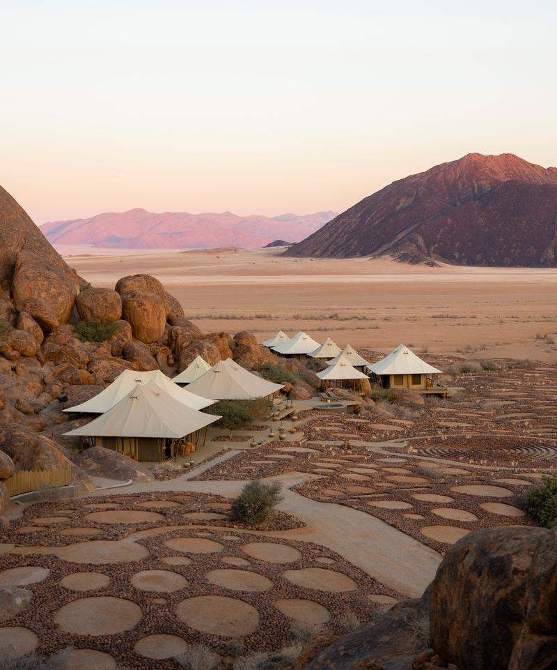 Cluster of white peaked tents at Wolwedans's Boulders Camp against large boulders in a wide desert plain.