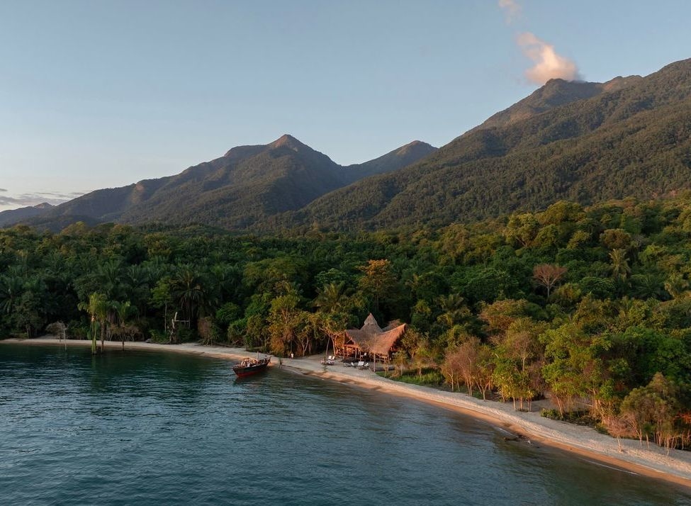 Aerial view of a remote beach lodge with thatched roofs on a white sand beach surrounded by dense jungle and mountains.