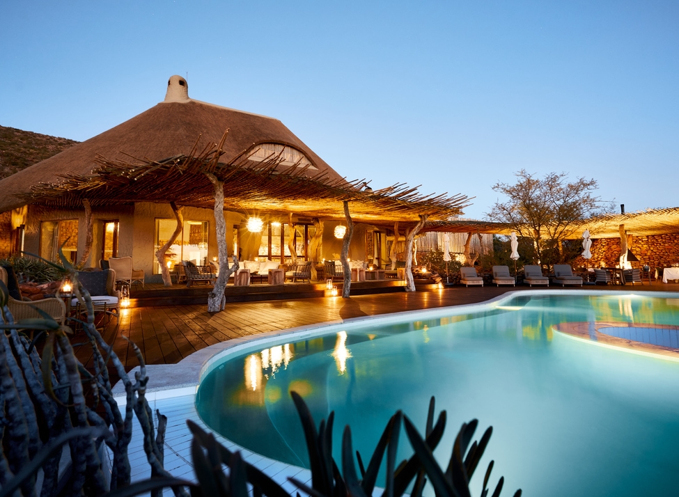A luxury safari lodge with a thatched roof and a glowing swimming pool on a wooden deck at dusk.