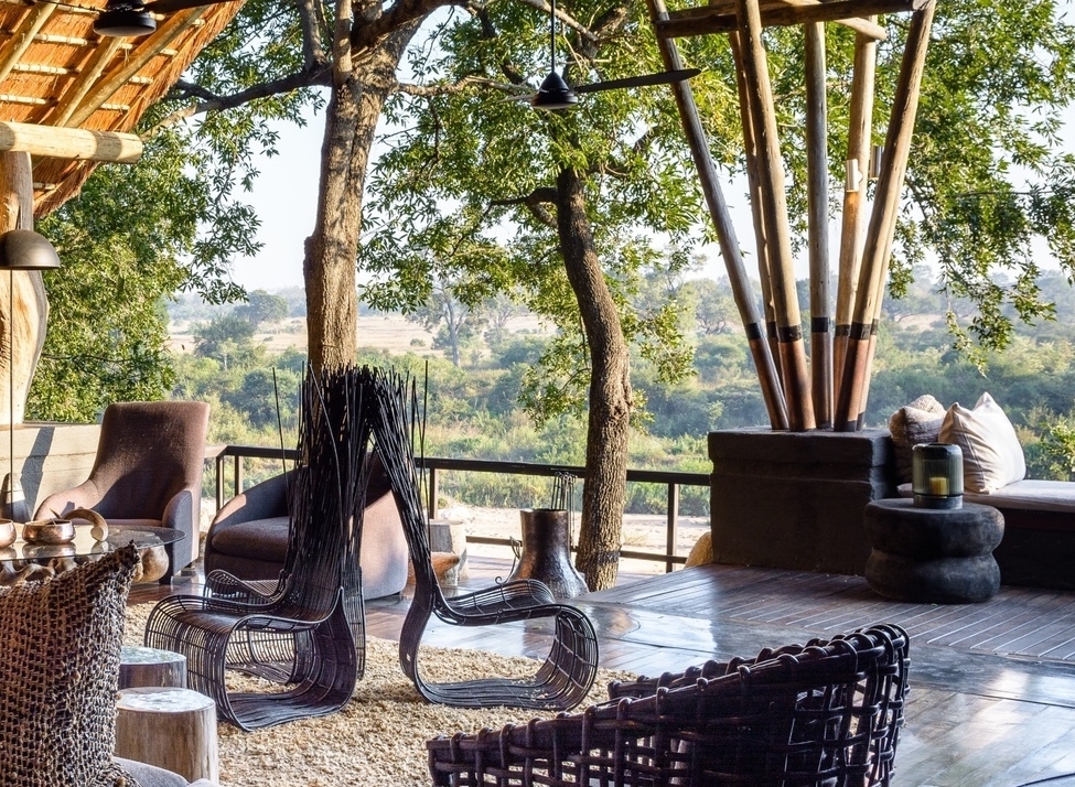 A luxury open-air lodge lounge with thatched roofing, woven chairs, and a scenic view of a riverbed and green trees.