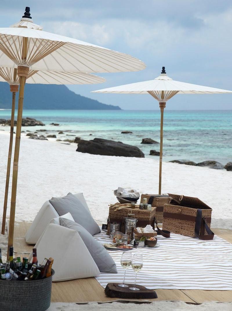 : A picnic set on a white sand beach with white umbrellas, cushions, food baskets and wine glasses.