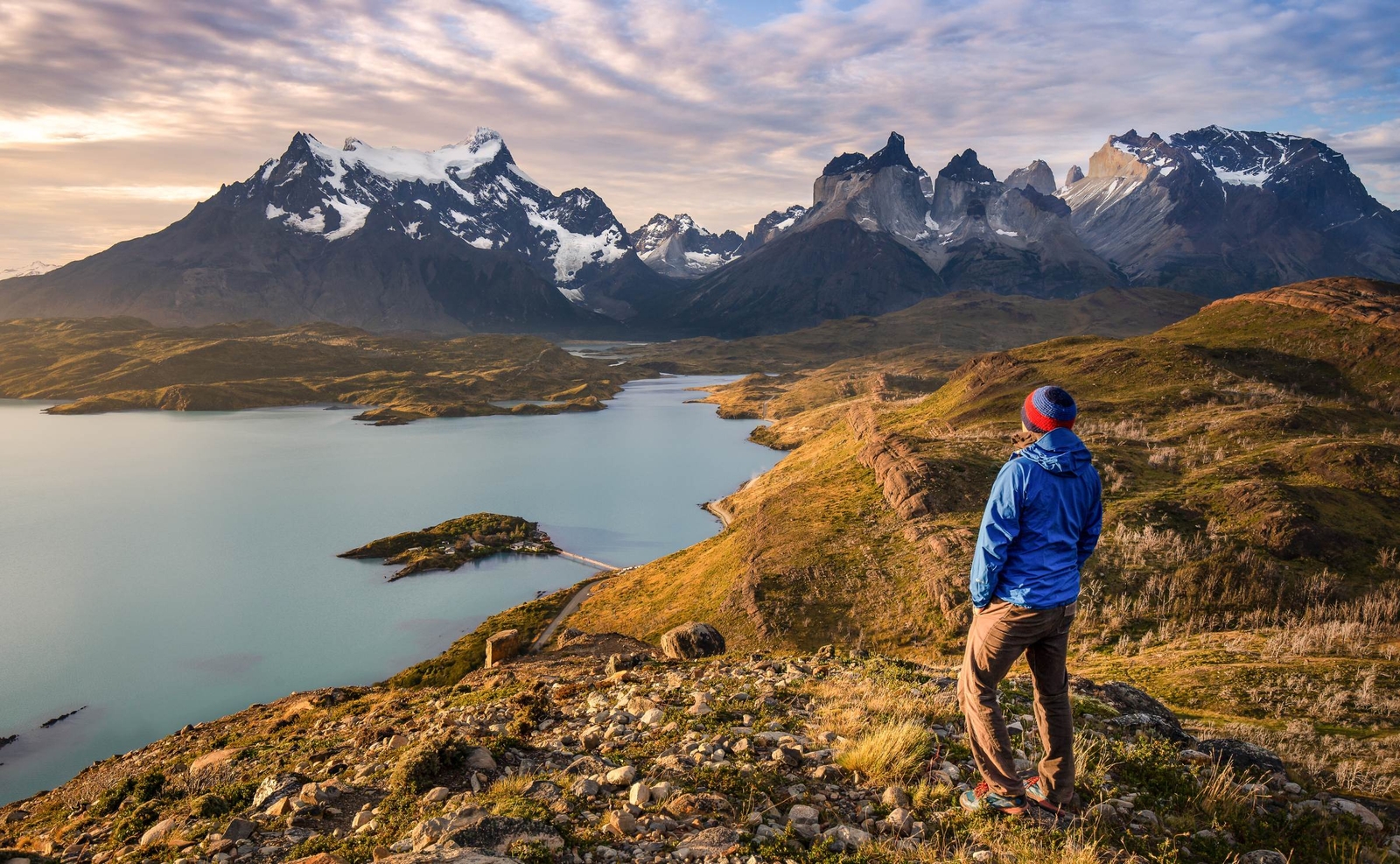 Back view of a person in a blue jacket standing on a hill looking at a wide lake and sharp mountain peaks.