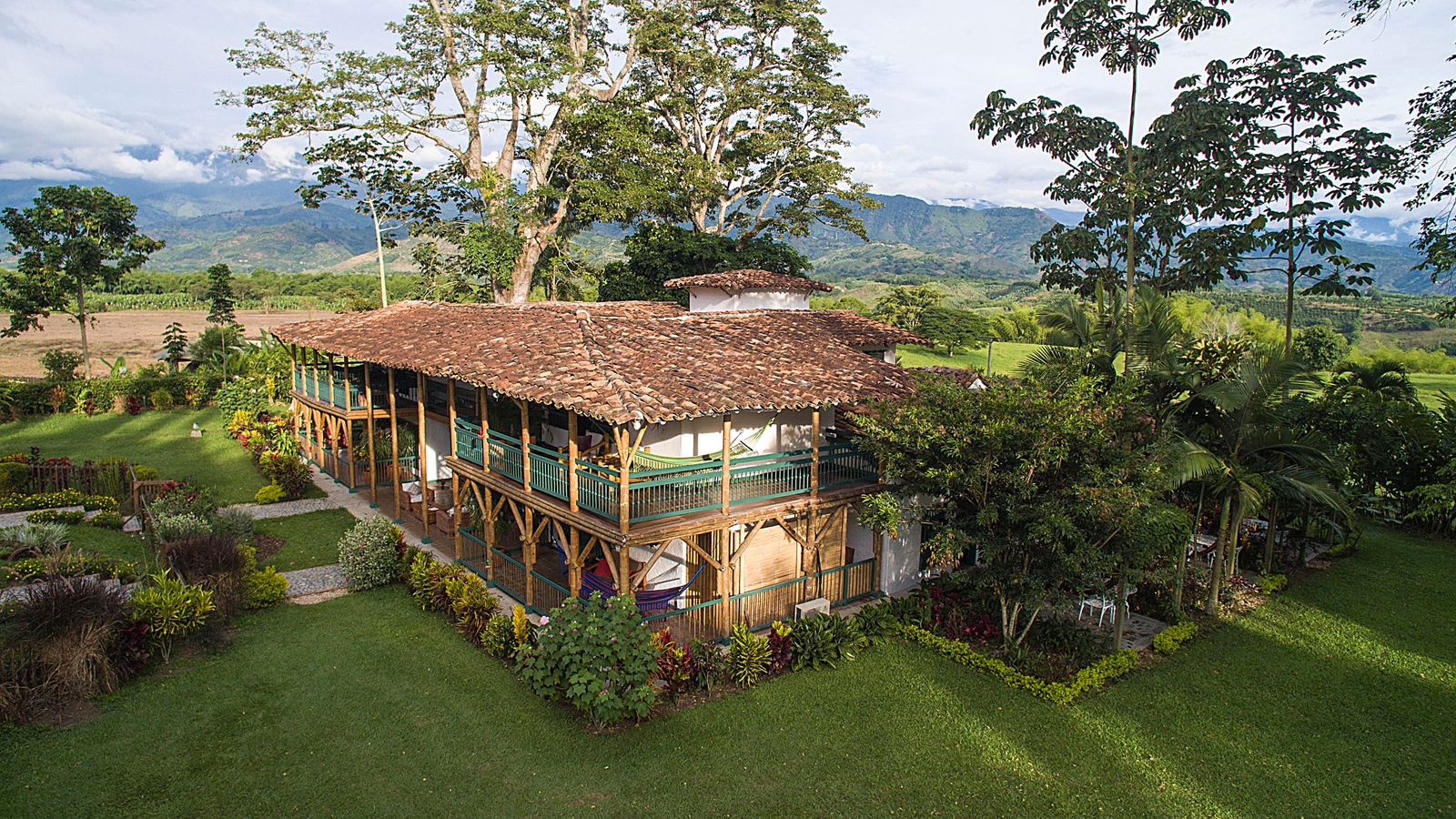 Large two-story wooden house with a balcony and tiled roof surrounded by green grass and distant mountains.
