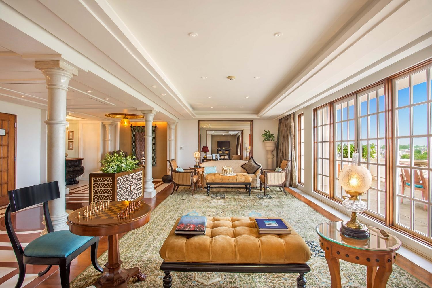 Elegant hotel living room with a chessboard, yellow tufted ottoman, and large windows overlooking a city.