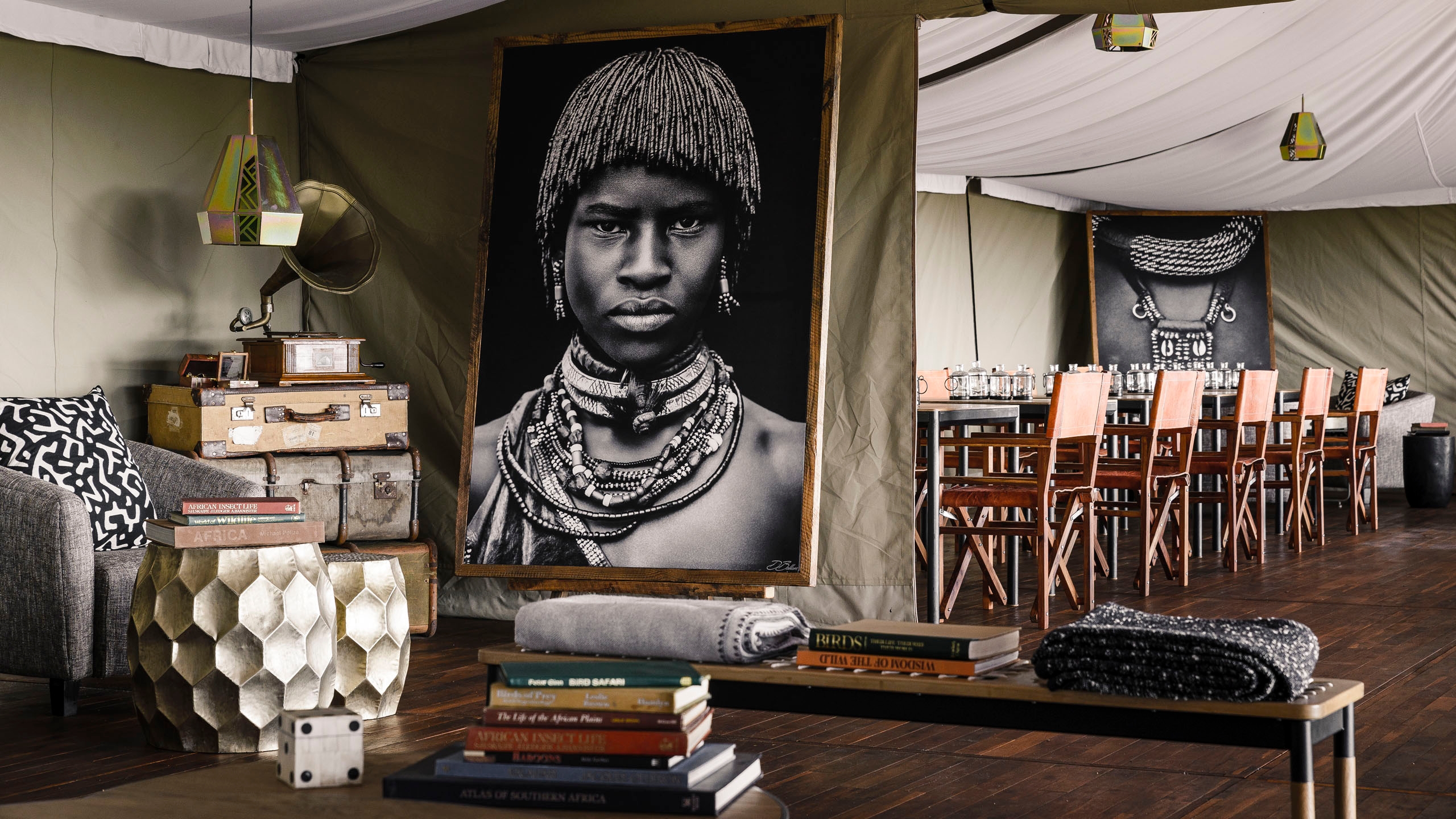 A covered dining room with a long table set with glasses and a living area with comfy chairs, a table and books, with black and white artwork on the walls