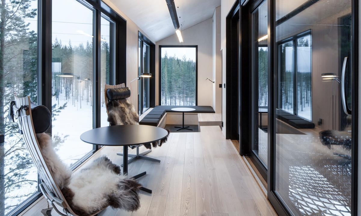 A modern cabin interior with light wood floors, large windows and chairs draped in thick animal furs.