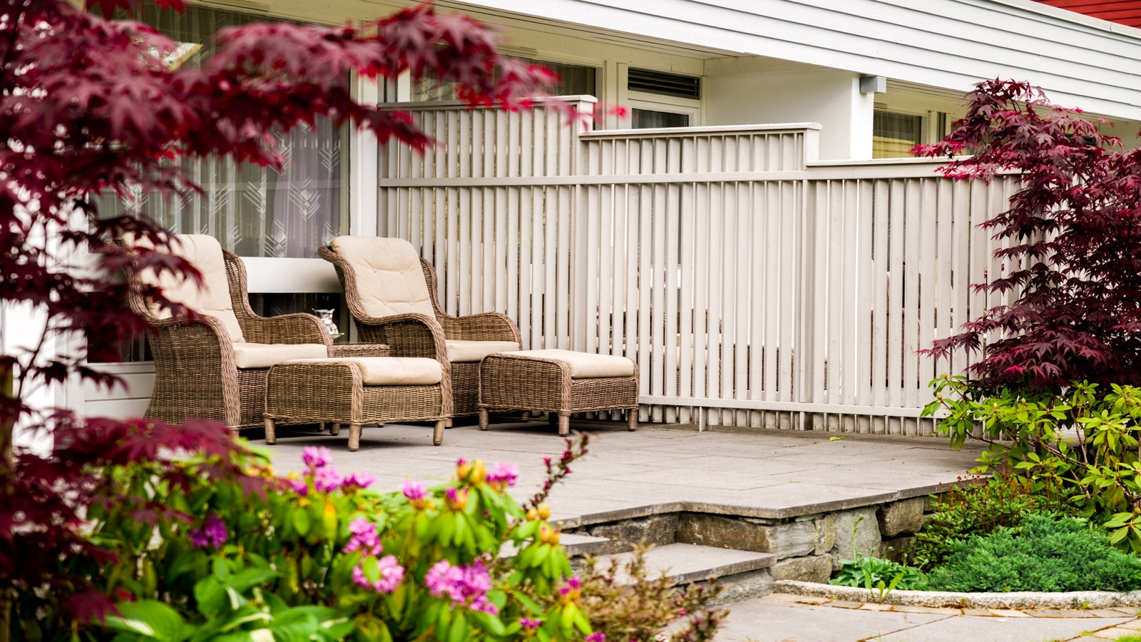 Cozy patio area with two wicker chairs, surrounded by lush greenery and red leaves.