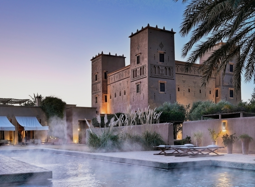 A steaming swimming pool at dusk with a large, lit-up Moroccan kasbah and palm trees in the background.