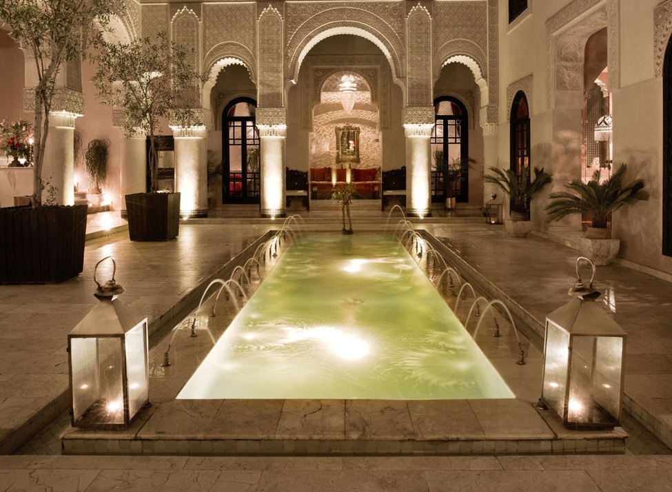 An ornate Moroccan courtyard at night with a glowing narrow pool, water fountains, and arched architecture.