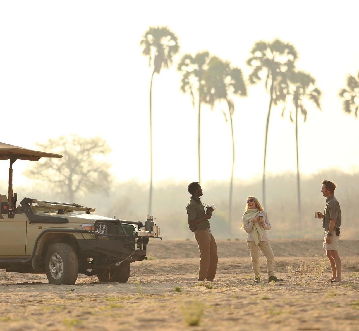 A couple and their guide enjoying beverages in the middle of the bush next to a safari vehicle.