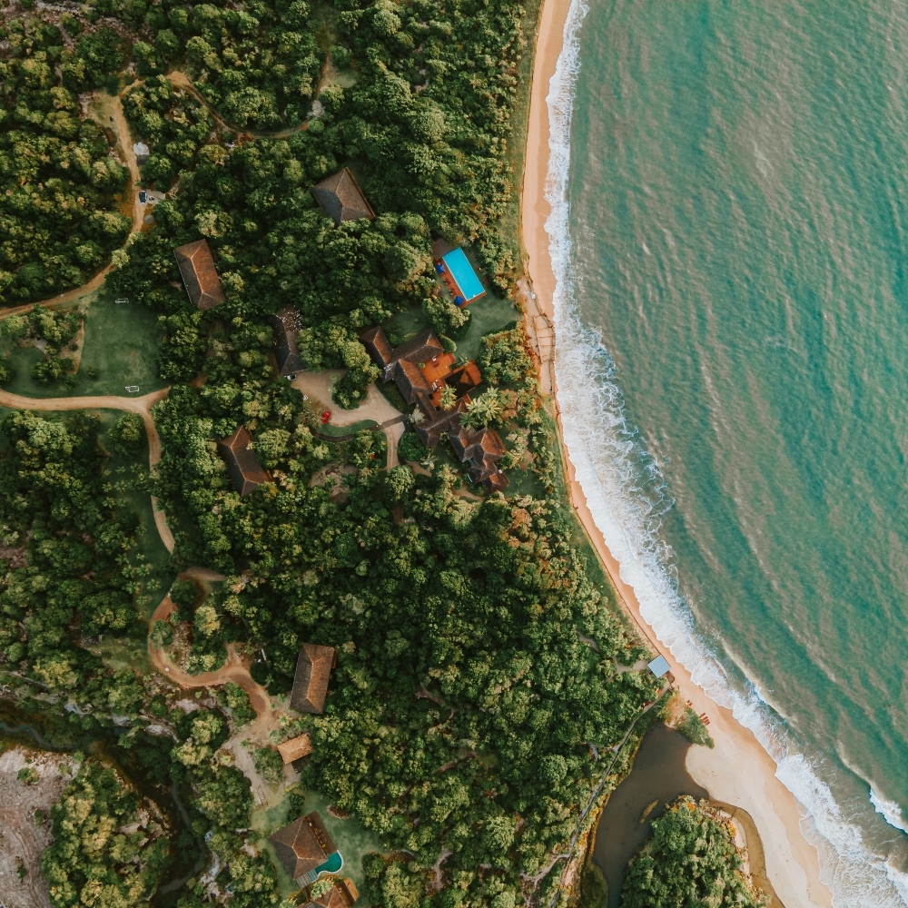 Aerial view of a lush coastline with a beach, ocean waves, and a pool amidst greenery.