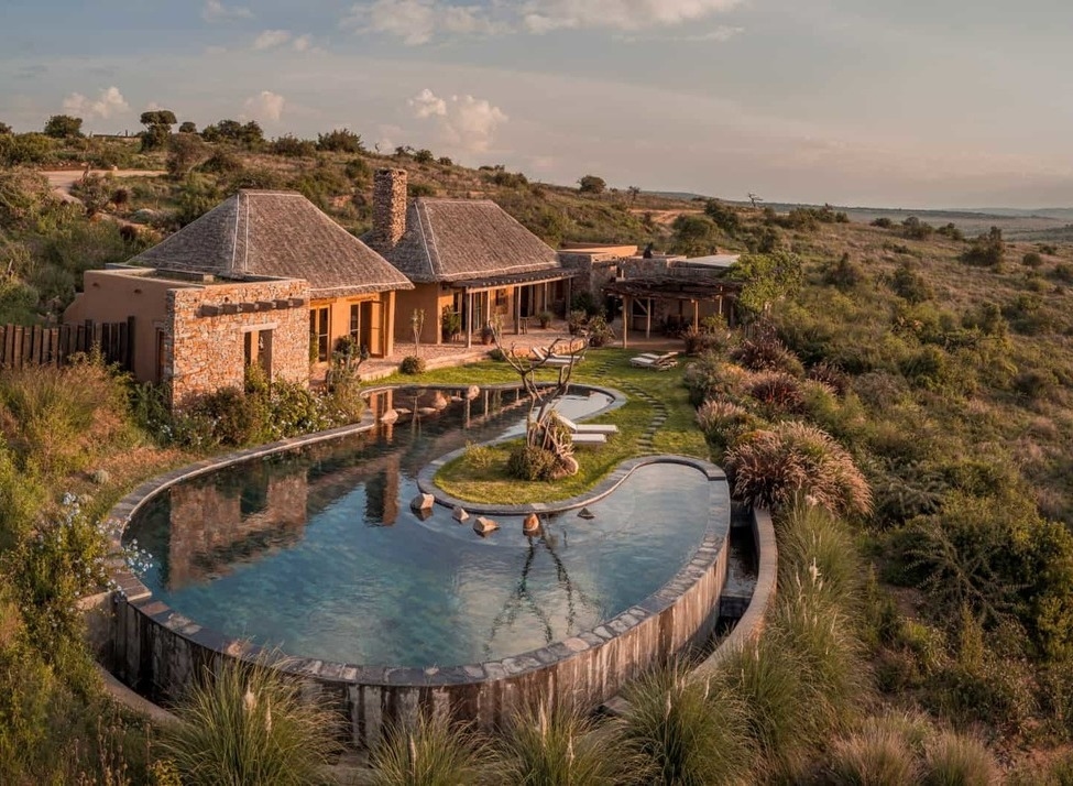 A stone luxury villa with a large, uniquely shaped infinity pool overlooking a wide, sunlit savanna landscape.