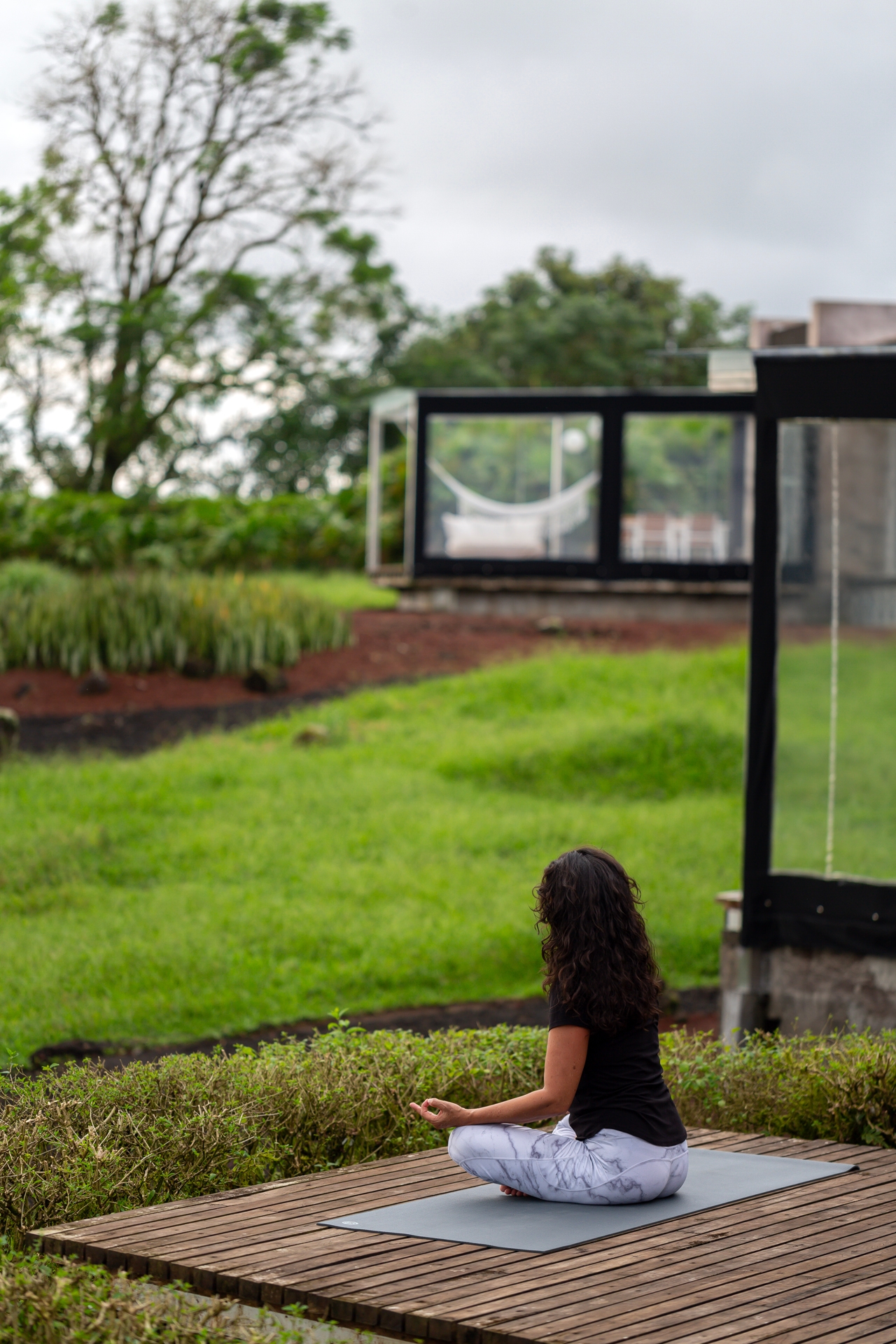 Woman meditating outdoors on a wooden deck with greenery and a modern structure in the background.