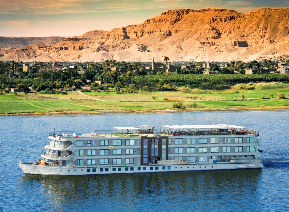 The Historia cruise ship sailing on the Nile River with green fields and arid mountains in the background.