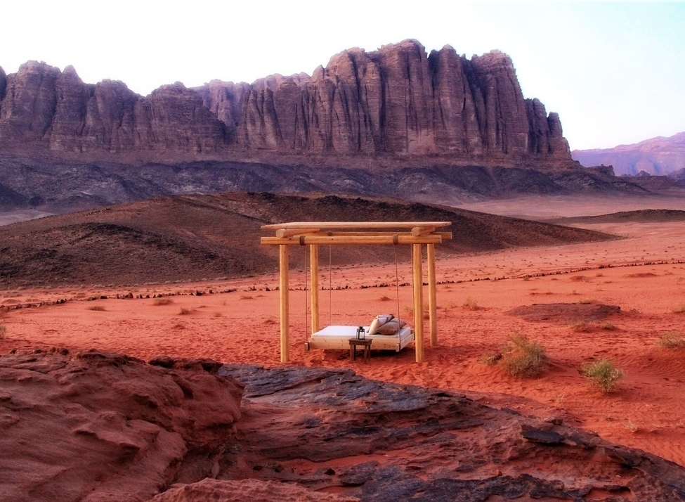 A wooden four-poster daybed on red desert sand with massive rock formations in the background at Discovery Bedu.