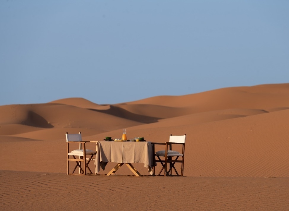 A breakfast table and two chairs set in the middle of vast desert sand dunes under a clear blue sky.