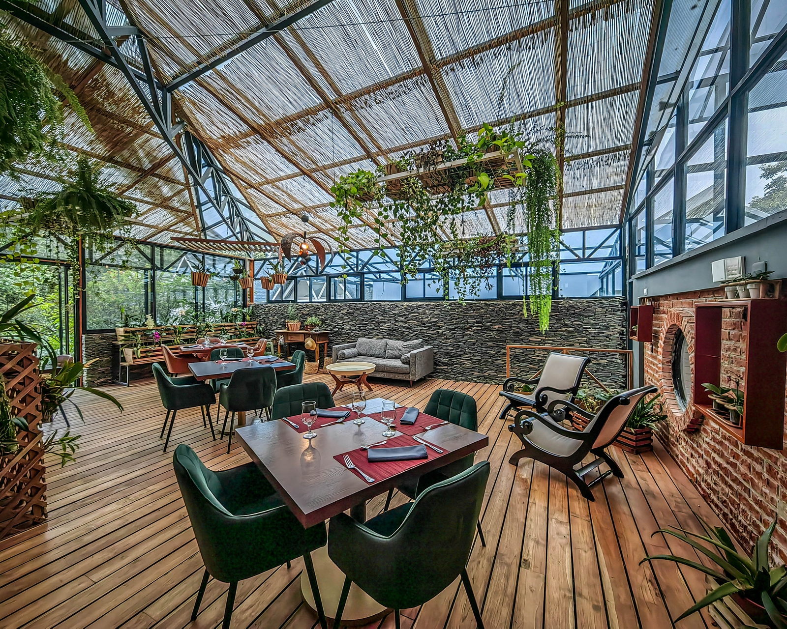 Indoor dining area with green chairs and wooden tables inside a glass-walled room filled with plants.