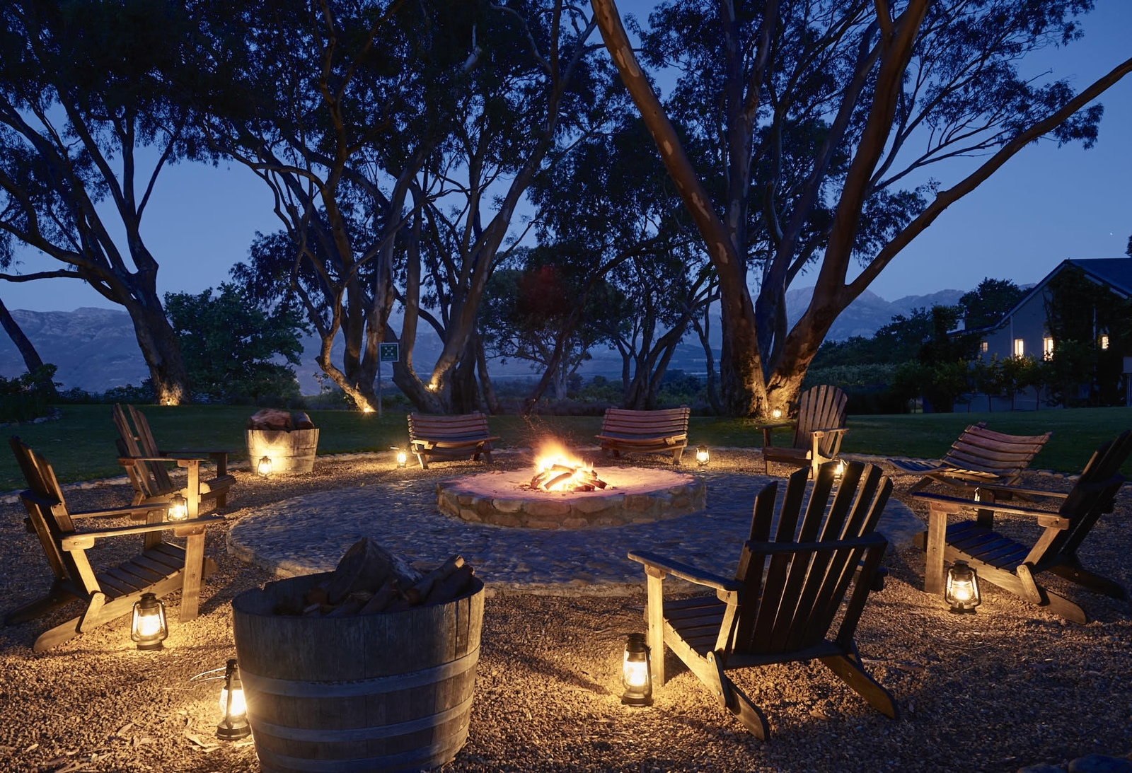 Outdoor fire pit surrounded by wooden chairs and lanterns at twilight, with trees and a house in the background.