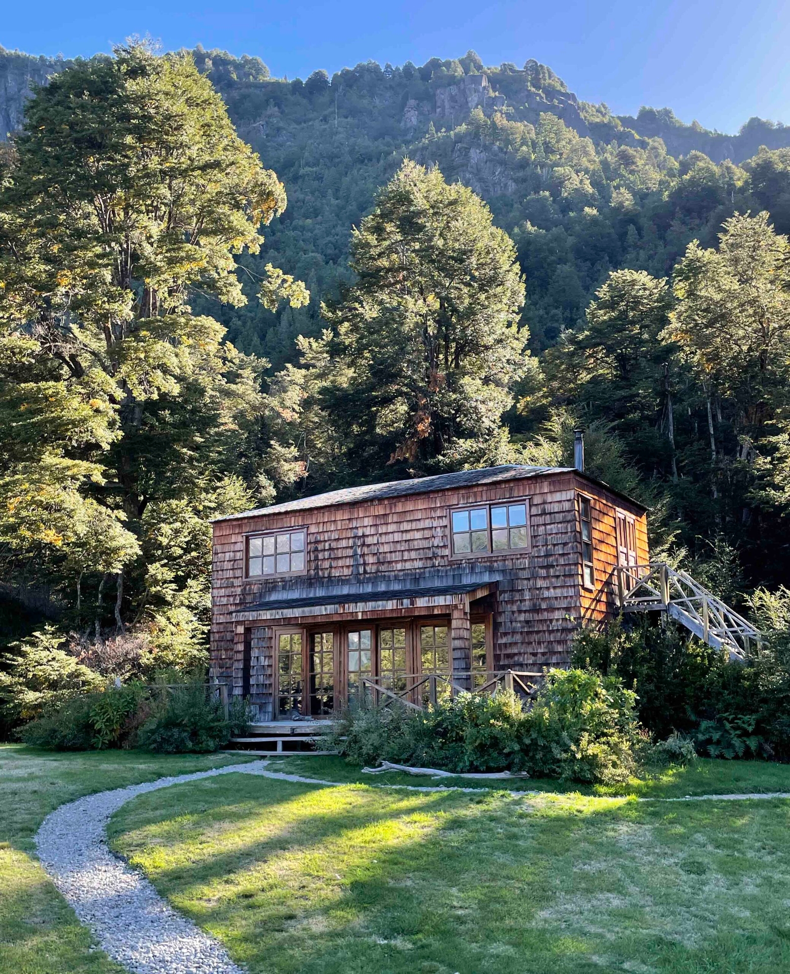 A wooden cabin surrounded by forest with a mountain backdrop under a clear sky.