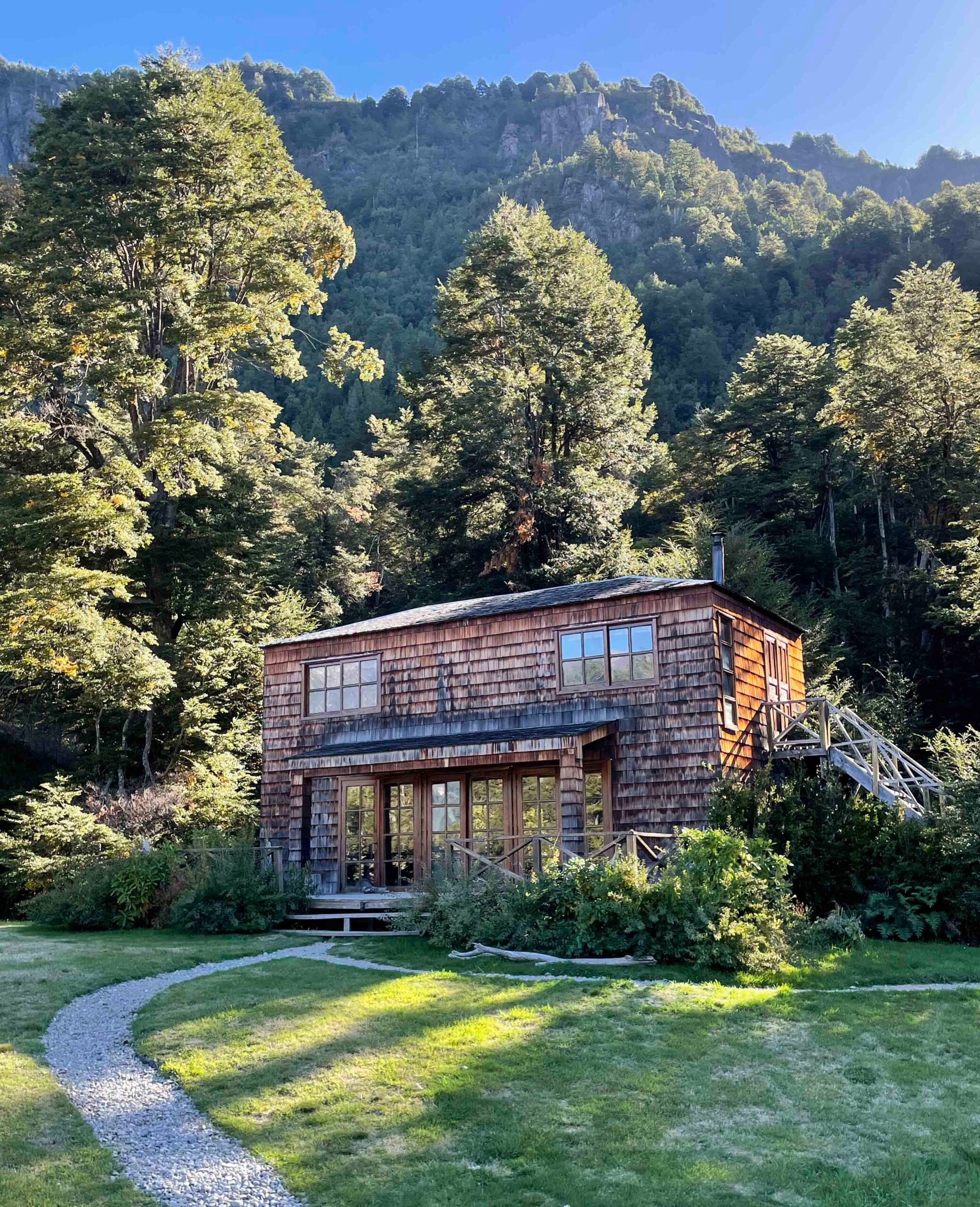 A wooden cabin surrounded by forest with a mountain backdrop under a clear sky.