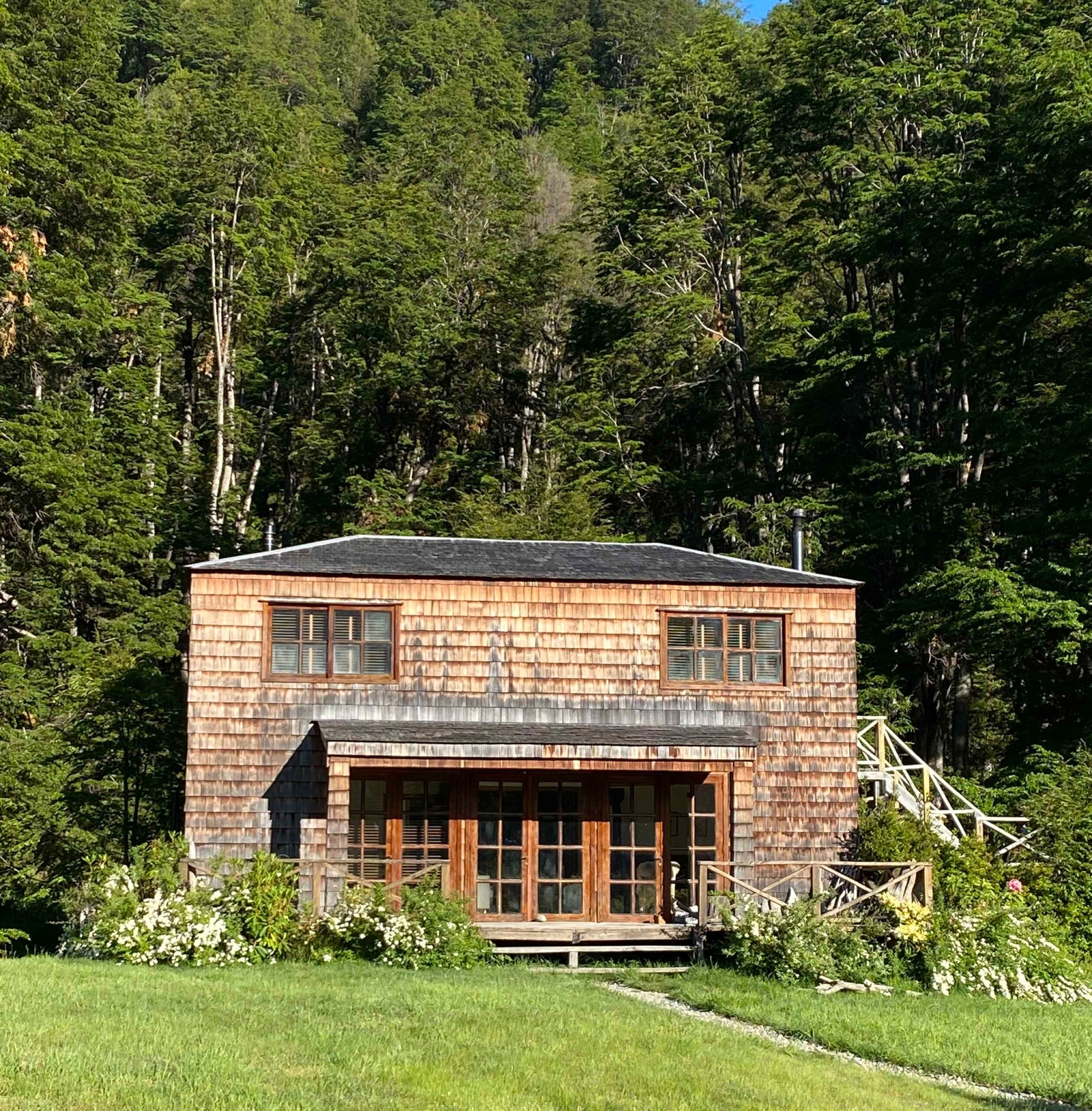 Wooden cabin with a shingled exterior in a clearing, surrounded by dense forest under clear skies.