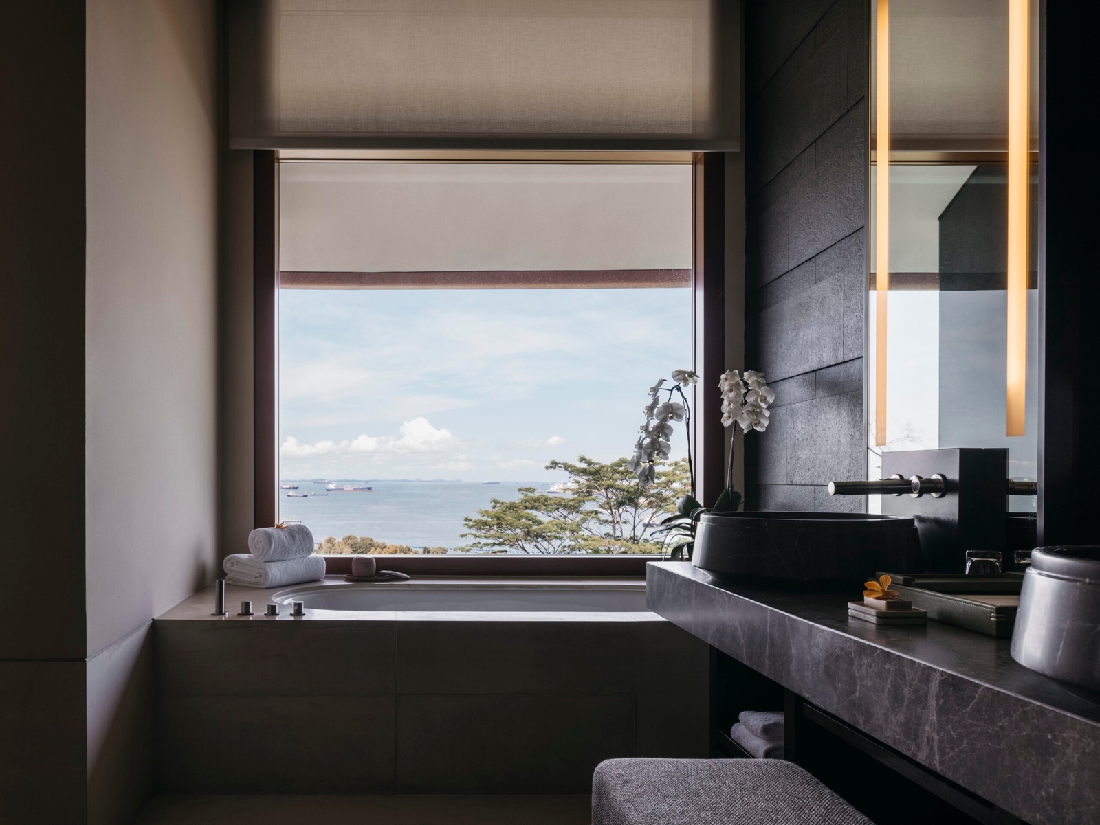 Modern bathroom with large window overlooking the ocean, featuring dark stone finishes and orchids.