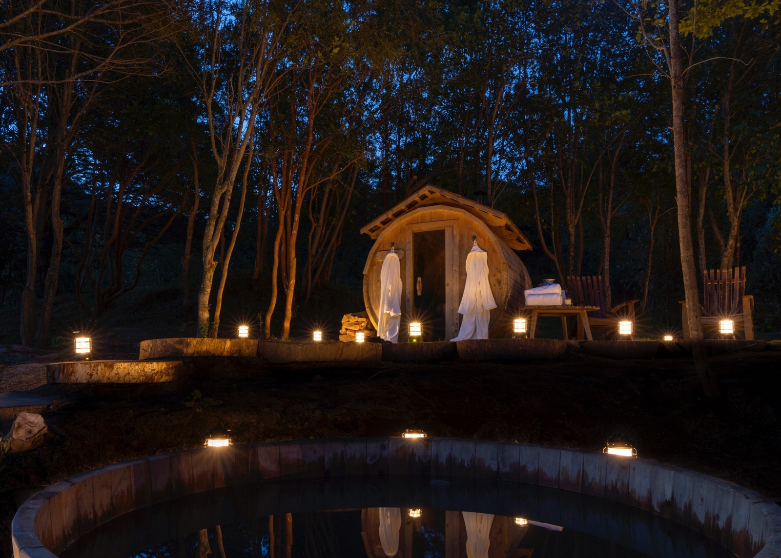 A cozy wooden sauna cabin by a pond, illuminated at dusk with lanterns.